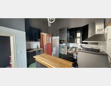 Moderne Küche mit schwarzen Hochglanzschränken, weißer Fliesenrückwand und zentraler Holzarbeitsinsel. Sichtbar sind Herd mit Edelstahl-Dunstabzug, Kühlschrank und Spüle; eine rote Innentür führt in den angrenzenden Bereich.