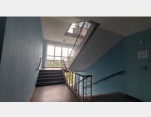 Treppe in einem modernen Gebäude mit blauen Wänden, Metallgeländern und einem großen rechteckigen Fenster, das Tageslicht hineinlässt und den Blick nach draußen ermöglicht. Die Treppe führt in verschiedene Stockwerke, und ein Fluchtplan ist an der Wand angebracht.