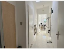 Schmaler, weiß gestrichener Flur mit Türen auf beiden Seiten, einem Kleiderständer rechts und einem Wandspiegel links; am Ende des Flurs ist ein heller Wohnraum mit einem Fenster und einer Sitzgelegenheit zu sehen.