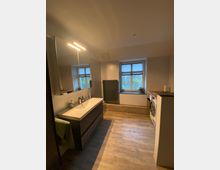 Modernes Badezimmer mit Holzoptik-Fliesenboden, einer hängenden Waschtischkombination und beleuchtetem Spiegel. Links befinden sich zwei Fenster mit halb geöffneten Jalousien, ein dunkler Heizkörper und eine Nische mit einer Waschmaschine.