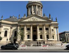 Außenansicht eines historischen Gebäudes mit klassizistischer Architektur, gekennzeichnet durch Säulen, Skulpturen und ein reich verziertes Giebeldreieck. Im Vordergrund sind eine Straße mit geparkten Autos, Absperrungen und Sitzplätze mit Sonnenschirmen auf einer Terrasse zu sehen.