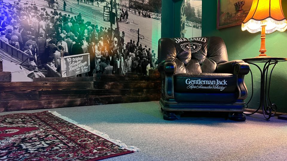 Gemütliche Lounge-Ecke mit grauem Teppich und gemustertem Teppichläufer, einem schwarzen Ledersessel mit Whiskey-Branding und einem kleinen Beistelltisch mit leuchtender Stehlampe. Im Hintergrund eine großflächige Schwarzweiß-Fototapete mit Straßenszene, dazu grün gestrichene Wände und ein gerahmtes Bild.