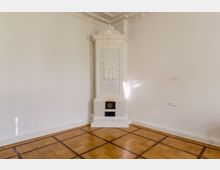 Heller Raum mit Parkettboden und weiß gestrichenen Wänden, in dem sich ein hoher, dekorativer Kachelofen mit Verzierungen in der Ecke befindet. Der Deckenrand ist mit Stuckornamenten dekoriert.