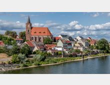 Kleinstadtansicht mit einer Backsteinkirche mit rotem Dach und einem hohen Turm im Zentrum, umgeben von Häusern mit bunten Dächern und Gärten. Im Vordergrund ein Fluss mit begrüntem Ufer, im Hintergrund Bäume und ein bewölkter Himmel.