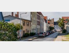 Straßenszene in einer Wohngegend mit mehrstöckigen, historischen Gebäuden, teilweise mit verzierten Fassaden und roten Ziegeldächern. Entlang der gepflasterten Fahrbahn sind parkende Autos zu sehen, und ein Straßenschild mit der Aufschrift „Karlstraße“ befindet sich im Vordergrund.