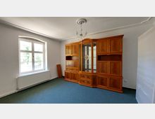 Ein leerer Raum mit weiß gestrichenen Wänden, blauem Teppichboden und einem großen, hölzernen Schrank mit verglasten und geschlossenen Fächern an der rechten Wand. Links befindet sich ein doppelflügeliges Fenster mit einem Heizkörper darunter.