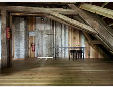 Holzvertäfelter Raum unter einem Dachstuhl mit sichtbaren Holzbalken. An der Rückwand stehen ein Tisch und mehrere gestapelte Hocker, daneben hängen ein Feuerlöscher und ein Gebäudeplan an der Wand.