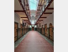 Korridor eines historischen Gefängnisses mit roten Böden, Holz- und Metalltüren zu den Zellen auf beiden Seiten und einem vergitterten Laufsteg im Obergeschoss. Oberlichtfenster und ein Netzstruktur an der Decke sind sichtbar.