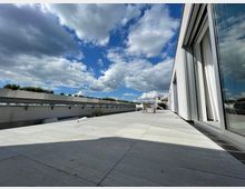 Weitläufige Terrasse mit hellen Steinfliesen, seitlichem Geländer mit Pflanzenbepflanzung und modernen Glasfronten auf der rechten Seite; im Hintergrund ein Tisch mit Stühlen unter teils bewölktem Himmel.