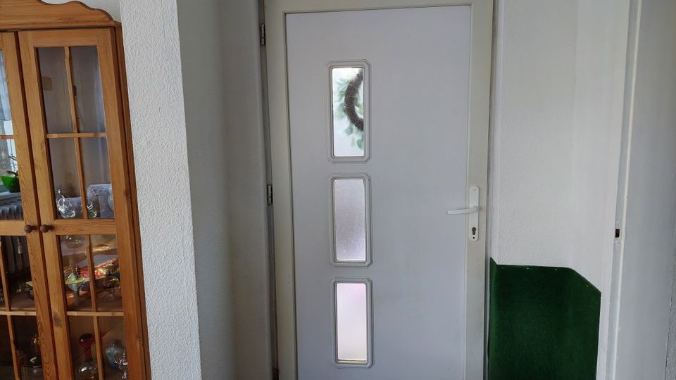 Innenflur mit einer weißen Eingangstür, die drei schmale, übereinander angeordnete Sichtfenster besitzt. Links steht eine hohe Vitrine aus Holz mit Glastüren, rechts ist ein grün gestrichener Wandbereich zu sehen.