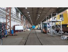 Ein Innenraum einer großen Werkshalle mit hohen Decken und Oberlichtern. Zu sehen sind Schienen auf dem Boden, ein gelber und ein blauer Zugwaggon, Regale mit Werkzeugen sowie ein Gabelstapler und metallene Arbeitsplattformen.