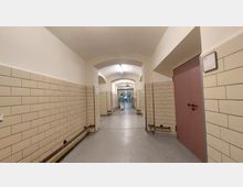 Langer Flur in einem Krankenhaus oder öffentlichen Gebäude mit cremefarbenen Fliesenwänden, gewölbter Decke und mehreren Türen; im Hintergrund geht eine Person in Arbeitskleidung.