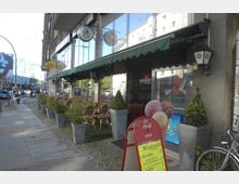 Straßencafé mit Glasfassade und grünem Vordach, ausgestattet mit kleinen Tischen und Korbstühlen auf einem Gehweg neben Pflanzenkübeln. Im Vordergrund steht ein Schild mit Speiseangeboten, und im Hintergrund sind Straßenverkehr sowie umliegende Gebäude zu erkennen.