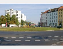Straßenkreuzung mit Zebrastreifen und einer begrünten Mittelinsel; im Hintergrund links mehrstöckige Plattenbauten, rechts historische Stadthäuser mit Geschäften im Erdgeschoss; ein U-Bahn-Schild markiert eine Station in der Mitte des Bildes.
