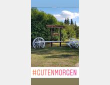 Holzschild mit der Inschrift „Gescht in Hammerbruch“ und „Einfahrt“, eingerahmt von zwei weißen Wagenrädern, auf einer Wiese vor dichter grüner Hecke und einigen Bäumen im Hintergrund. Der Himmel ist blau mit weißen Wolken.