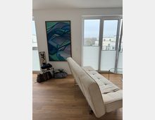 Helles Wohnzimmer mit Parkettboden, einem beigen Sofa, einer großen Fensterfront mit weißen Plissees und einem abstrakten Gemälde an der Wand. Im Vordergrund stehen mehrere Taschen und ein kleiner Hocker.