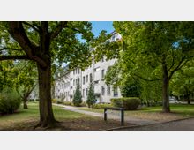 Mehrstöckiges Wohngebäude mit weißer Fassade und blauen Fensterrahmen, umgeben von Grünflächen mit Bäumen und Gehwegen. Eine Hausnummertafel steht am Rand des Gehwegs im Vordergrund, und die Fassade eines Teils des Gebäudes ist mit Efeu bewachsen.