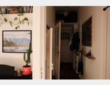 Schmaler Wohnungsflur mit halb geöffneter Tür und Blick zur Eingangstür am Ende des Gangs. An der rechten Wand hängen Garderobenhaken, ein Foto-Board und ein schmales Regal; links sind ein gerahmtes Landschaftsbild, Zimmerpflanzen und ein kleines Sideboard zu sehen.