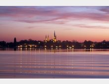 Eine ruhige Seelandschaft bei Sonnenuntergang mit beleuchteten Gebäuden im Hintergrund, unter anderem eine Kirche mit zwei hohen Türmen. Die Beleuchtung spiegelt sich im Wasser, das von sanften Wellen übersät ist.