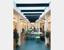 Ein moderner Innenraum mit Sitzbereichen und Arbeitsflächen, gestaltet als Gemeinschafts- oder Arbeitsbereich. Der Raum hat eine Glasdecke, weiße Säulen, eine offene Gestaltung und ist mit Pflanzen sowie schlichten Stühlen und Tischen eingerichtet.