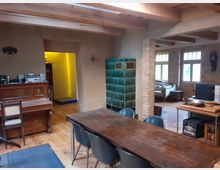 Wohnraum in rustikalem Stil mit Holzboden, freiliegenden Holzbalken und Backsteinwänden. Zu sehen sind ein grüner Kachelofen, ein Holztisch mit Stühlen, ein Klavier, sowie ein Sitzbereich mit Sofas im Hintergrund.