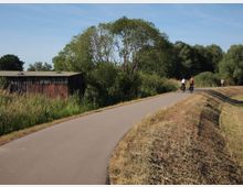 Eine asphaltierte Rad- und Fußwegstrecke führt durch eine grüne Landschaft mit Bäumen und Büschen. Links steht ein altes, hölzernes Gebäude neben Schilf, während mehrere Personen auf Fahrrädern und zu Fuß den Weg nutzen.