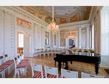 Prunkvoller Innenraum eines historischen Veranstaltungsraums mit Stuckverzierungen, großen Kristallkronleuchtern, bemalter Decke und weißen Wänden. Im Vordergrund ein schwarzer Flügel, umgeben von weiß-roten Stühlen auf einem Parkettboden.