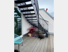 Eine Dachterrasse mit Holzboden und einer schwarzen Metalltreppe, die nach oben führt. Auf der Terrasse stehen ein türkisfarbener Stuhl, ein roter Klapptisch mit passendem Stuhl sowie mehrere Blumentöpfe, darunter ein großer, runder Pflanzkübel.