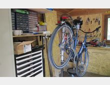 Eine Werkstatt mit Holzwänden, in der ein Fahrrad auf einem Reparaturständer befestigt ist. Im Hintergrund sind Werkzeuge an einer Wandhalterung angebracht, daneben befinden sich eine Werkbank und Schubladenschränke mit Kleinteilesortiment.