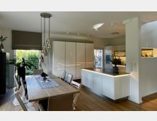 Offener Wohn- und Essbereich mit moderner, weißer Einbauküche und Kücheninsel mit dunkler Arbeitsplatte sowie indirekter Beleuchtung. Daneben steht ein großer Holzesstisch mit Stühlen unter hängenden Pendelleuchten, mit großen Fenstern und Blick ins Grüne.