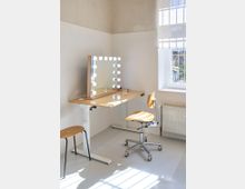 Ein heller Raum mit einem höhenverstellbaren Holztisch, darauf ein Spiegel mit runden Leuchten, ein drehbarer Holzstuhl auf Rollen und ein zusätzlicher Hocker. Rechts im Bild befindet sich ein Fenster mit einem halbhoch heruntergelassenen Rollo und darunter ein Heizkörper.