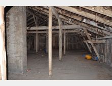 Dachbodenraum eines alten Gebäudes mit sichtbaren hölzernen Dachbalken, Holzstützen und einem unebenen Holzboden. Die Wände sind roh, und im Raum stehen vereinzelte Gegenstände wie Eimer und Leitern.