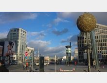 Straßenszene am Potsdamer Platz in Berlin mit modernen Gebäuden, einem Uhrenturm und Werbetafeln; im Hintergrund ist der Berliner Fernsehturm sichtbar. Auf einem Laternenmast hängt eine große goldene Kugel als Dekoration.