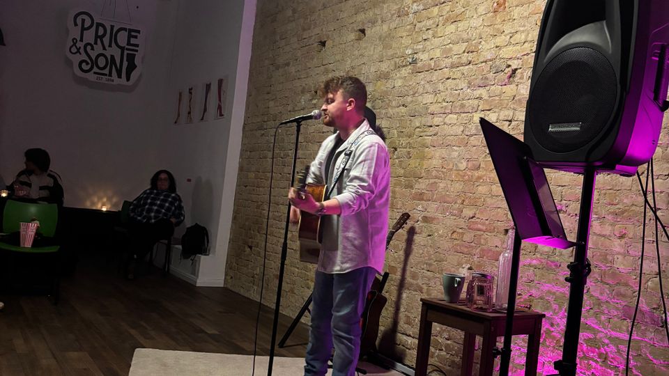Small indoor performance space with an exposed brick wall, a rug-covered stage area, and purple accent lighting. A microphone stand, speaker on a tripod, music stand, and a stool with equipment sit beside the stage, with seating and a “Price & Son” sign visible at the back.