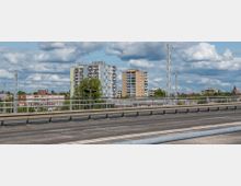 Straßenansicht mit Geländer im Vordergrund, dahinter mehrere farbige Wohnhochhäuser sowie ein Kirchturm in einer städtischen Umgebung, unter einem bewölkten Himmel. Im Hintergrund sind Strommasten und weitere Gebäude sichtbar.