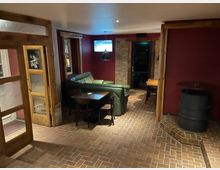 Gemütlicher Innenraum eines Lokals mit roten Wänden und Backsteinboden. Zu sehen sind Ledersofas, Holzmöbel, eine Holztür mit Glasfenstern sowie eine Wand mit einem Fernseher darüber.