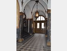 Eingangsbereich eines historischen Gebäudes mit Verzierungen, einschließlich marmorgleicher Säulen und goldener Details, gewölbten Decken, einer hölzernen Doppeltür mit milchglasartigen Fenstern, einer großen Laternen-Hängeleuchte und einer Treppe auf der linken Seite. Der Boden ist mit einem geometrischen Fliesenmuster gestaltet.