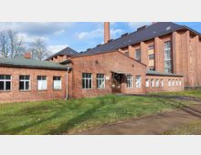 Backsteingebäude mit einfachem Satteldach und zahlreichen rechteckigen Fenstern, ergänzt durch einen größeren Anbau mit modernen Fenstern und einem Schornstein im Hintergrund. Das Gebäude steht auf einer Rasenfläche, umgeben von kahlen Bäumen und Gehwegen.