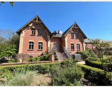 Backsteinvilla im historischen Stil mit zwei symmetrischen Giebeldächern und dekorativen Holzverzierungen. Eine Freitreppe führt zur Eingangstür, umgeben von einem gepflegten Garten mit Hecken, Sträuchern und Blumen.