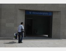 Eingang zur U-Bahn-Station "Alexanderplatz" mit einem blauen Schild über der Tür. Eine Person mit Einkaufstüten und einer Handtasche geht auf die Station zu.