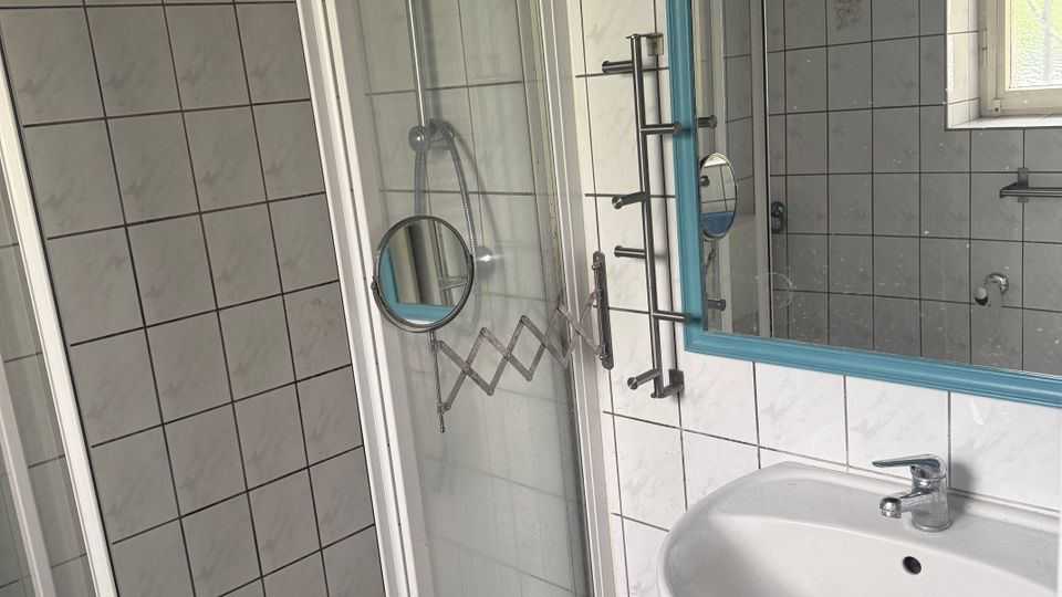 Kleines, gefliestes Badezimmer mit Duschwanne und Glas-Schiebetür sowie einem Waschbecken neben einem großen Spiegel mit blauem Rahmen. Rechts sorgt ein Fenster für Tageslicht; die Wände und der Boden sind mit weißen Fliesen im Rastermuster versehen.