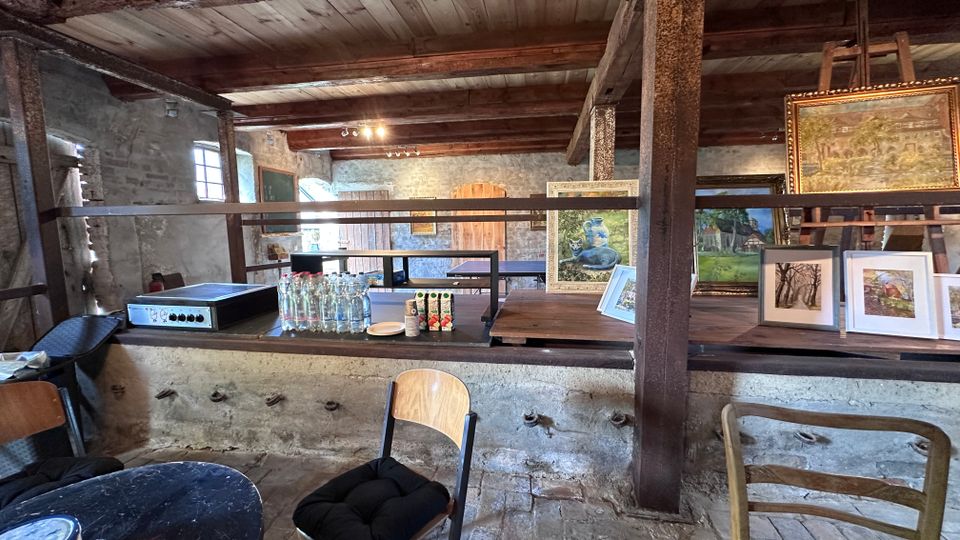 Innenraum eines rustikalen Steingebäudes mit sichtbaren Holzbalken und Holzdecke, in dem auf einem langen Tresen gerahmte Gemälde und Drucke ausgestellt sind. Im Vordergrund stehen Stühle und ein Tisch, daneben befinden sich Flaschen und Gläser auf dem Tresen; kleine Fenster und Holztüren prägen den Raum.