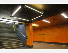 U-Bahn-Station mit orangefarbenen Wandverkleidungen und grauen Fliesen an den Treppenwänden. Die Decke ist mit schwarzen und weißen Leuchtstoffröhren beleuchtet, und ein Schild weist zur Treppe mit der Aufschrift „Ausgang“.