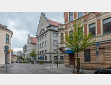 Straßenszene in einer urbanen Umgebung mit historischen Gebäuden. Mehrstöckige Gebäude mit Satteldächern und verzierten Fassaden grenzen an eine Kopfsteinpflasterstraße, flankiert von geparkten Autos und kleinen Bäumen.