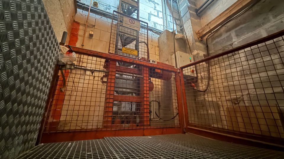 Innenansicht eines industriellen Technik- oder Aufzugsschachts mit Metallgitterboden und rotbraunen Schutzgittern. Sichtbar sind Betonwände, Rohrleitungen, Kabel, ein Schaltschrank und eine vertikale Stahlkonstruktion mit Warnschildern.