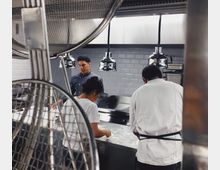 Drei Personen arbeiten in einer modernen Profiküche mit Edelstahloberflächen und Hängelampen. Im Vordergrund sind Küchenutensilien sichtbar, während weiße Fliesenwände den Hintergrund bilden.