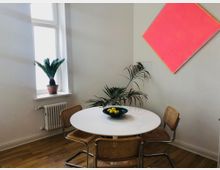 Ein minimalistisch eingerichteter Raum mit einem runden weißen Esstisch, umgeben von vier Holzstühlen mit geflochtenen Sitzflächen. Im Hintergrund befinden sich ein großes Fenster mit einem kleinen Palmengewächs auf der Fensterbank, ein Heizkörper darunter und eine Wand mit einem pinkfarbenen, schräg aufgehängten Gemälde.