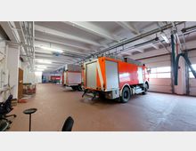 Eine Feuerwehrgarage mit geparkten roten Einsatzfahrzeugen, großer Deckenbeleuchtung, Lüftungsrohren und Rolltoren im Hintergrund. Der Boden besteht aus roten Fliesen, und an den Seiten stehen verschiedene Ausrüstungsgegenstände.