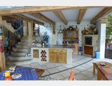 Offene Küche im rustikalen Stil mit freiliegenden Holzbalken an der Decke und dekorativen Natursteinelementen an den Wänden. Sichtbar sind eine Kücheninsel mit Holzschränken, eine gewundene Steintreppe, offene Regale und ein Backofen in einer gemauerten Nische.