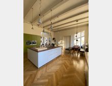 Geräumige, modern eingerichtete Küche mit einer zentralen Kücheninsel, weißen Schränken und einer Holzarbeitsplatte, einem hölzernen Fischgrät-Parkettboden, einer grünen Einbauschrankwand und mehreren Pendelleuchten an der Decke. Im Hintergrund ein Essbereich mit einem Holztisch, Bänken und großen Fenstern, die Tageslicht hereinlassen.